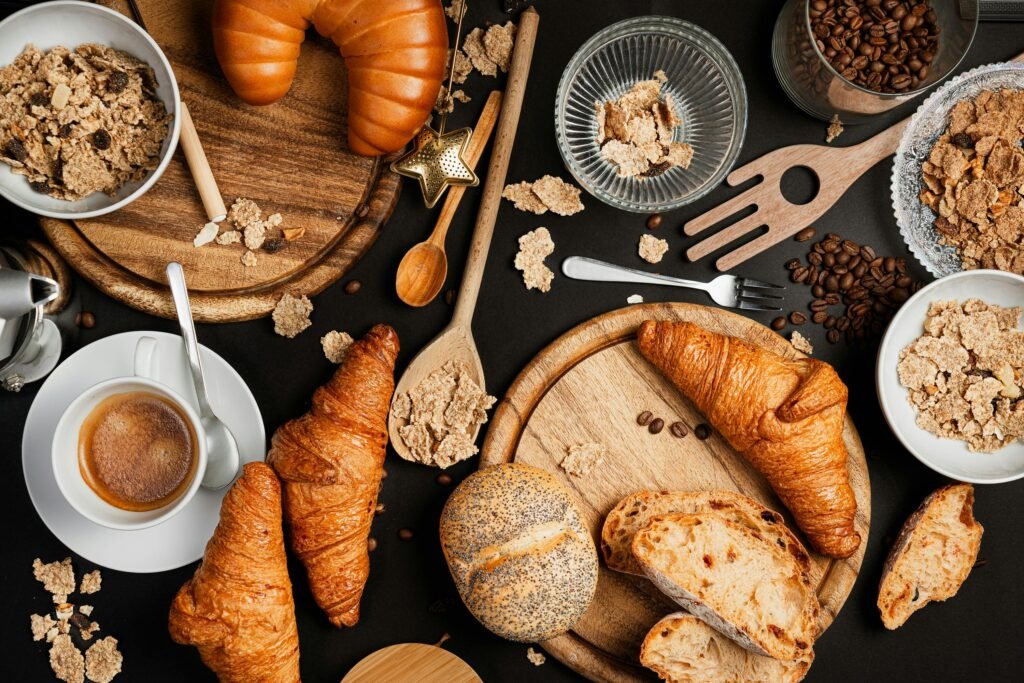 Top view of a breakfast spread featuring croissants, breads, cereals, and coffee on a table.